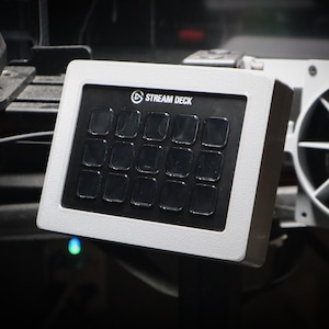 May include: A white Stream Deck with 15 black buttons. The buttons are arranged in a 5x3 grid. The Stream Deck is a device that allows users to control their streaming software with the push of a button.