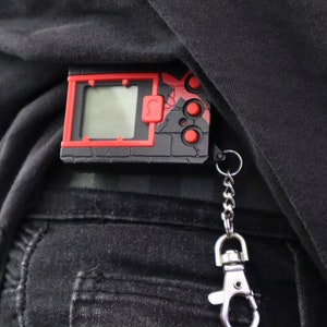 May include: A black and red digital device with a screen, buttons, and a keychain attachment. The device is clipped to a black garment. The design features a red border and accents, with a small screen displaying a digital interface.