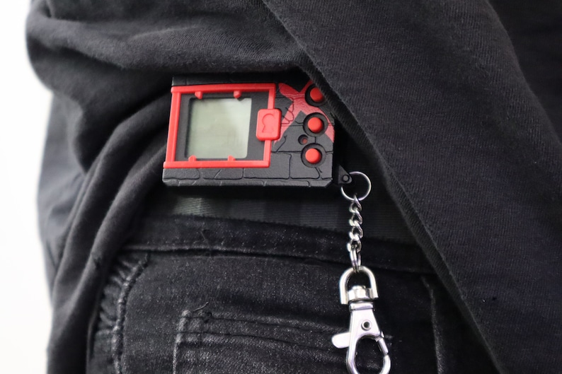 May include: A black and red digital device with a screen, buttons, and a keychain attachment. The device is clipped to a black garment. The design features a red border and accents, with a small screen displaying a digital interface.