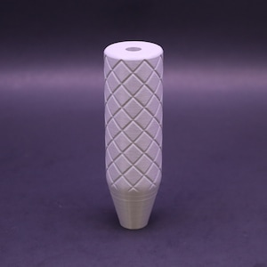 May include: A light grey, cylindrical object with a diamond pattern. The top has a circular opening. The bottom tapers to a point. It is likely a decorative or functional item, possibly a handle or knob.