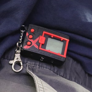 May include: A black and red handheld digital device with a small screen and buttons, attached to a silver keychain. The device has a red X design and is resting on a dark blue fabric.