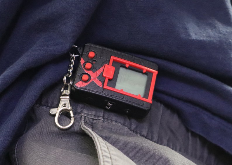 May include: A black and red handheld digital device with a small screen and buttons, attached to a silver keychain. The device has a red X design and is resting on a dark blue fabric.