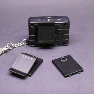 May include: A black handheld digital device with a silver chain and clasp. The device has a rectangular screen and several buttons. Two black rectangular covers and a black cover with a circular hole are also present.