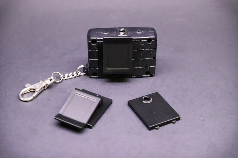 May include: A black handheld digital device with a silver chain and clasp. The device has a rectangular screen and several buttons. Two black rectangular covers and a black cover with a circular hole are also present.