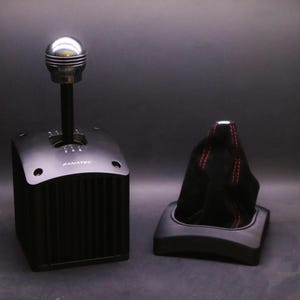 Könnte beinhalten: Ein schwarzer und silberner Rennsimulator-Schalthebel mit einem Metall-Schaltknauf. Auf der Basis steht die Marke "FANATEC". Daneben befindet sich eine schwarze Schalthebelmanschette mit roten Nähten.