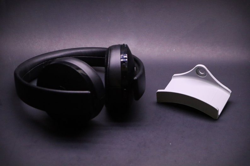 May include: A black pair of over-ear headphones with a white stand. The headphones are lying on a dark surface.