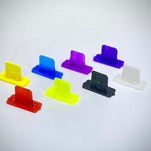 Może przedstawiać: Kolekcja ośmiu małych, prostokątnych plastikowych przedmiotów w różnych kolorach, w tym żółtym, niebieskim, fioletowym, czarnym i białym. Każdy przedmiot ma podniesioną, prostokątną sekcję na górze. Przedmioty są ułożone na białej powierzchni.