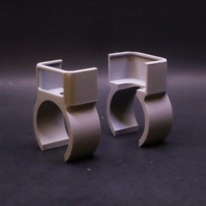 May include: Two light gray, 3D-printed plastic clips. Each clip has a rounded base and a rectangular top section. The clips are designed to hold something, with a curved opening in the center. The background is a dark gray.