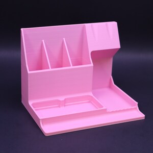 May include: A pink plastic desk organizer with four compartments, a small tray, and a larger tray. The organizer is designed to hold pens, pencils, and other office supplies.