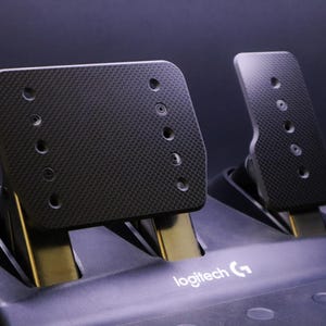 Puede incluir: Primer plano de un juego de pedales de carreras Logitech G negros. Los pedales tienen una superficie texturizada con múltiples agujeros. La base es negra con el logotipo de Logitech G.