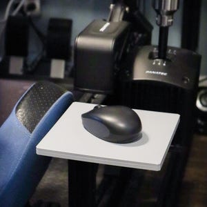 May include: A black computer mouse with gray accents sits on a white rectangular platform. The platform is part of a black gaming setup, with a blue footrest visible on the left. Other black components are in the background.