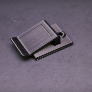 May include: A black, rectangular clip with a hinged top, designed to hold items. The clip has a textured surface and a small hole on the base. The object is made of a dark material.