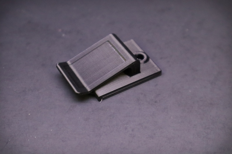 May include: A black, rectangular clip with a hinged top, designed to hold items. The clip has a textured surface and a small hole on the base. The object is made of a dark material.
