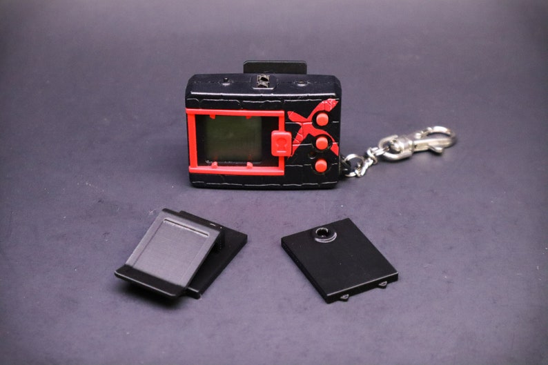 May include: A black and red handheld digital device with a small screen, buttons, and a keychain attachment. The device has a red border around the screen and red accents. Two black battery covers are next to the device.