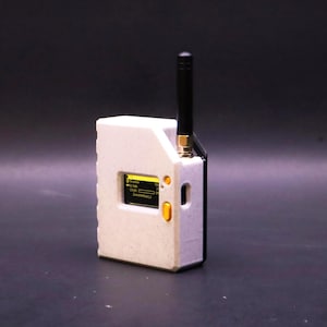 以下が含まれることがあります： 白いテクスチャ加工が施された長方形の電子機器で、黒い背面パネルと垂直な黒いアンテナを備えています。前面には黄色い文字が表示される小さなデジタル画面があり、ケースの右側には2つの黄色い円形ボタンが配置されています。