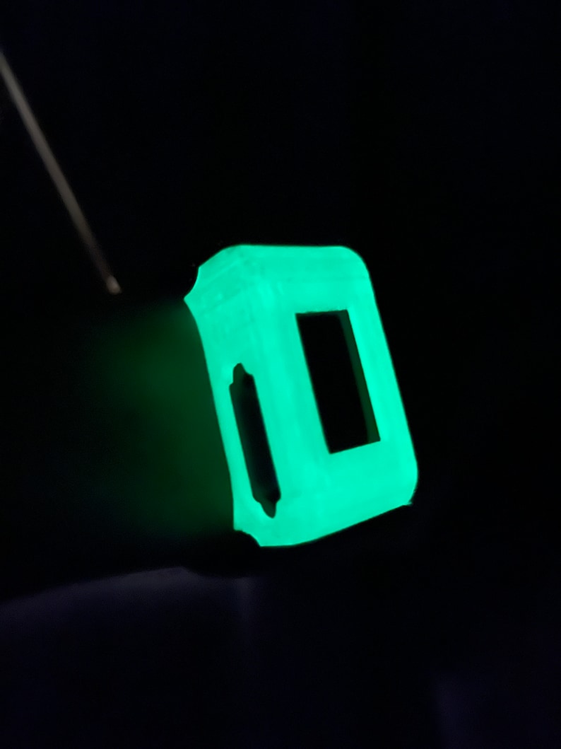 Pode incluir: Um pequeno objeto quadrado feito de um material que brilha no escuro. O objeto tem uma abertura retangular no centro.