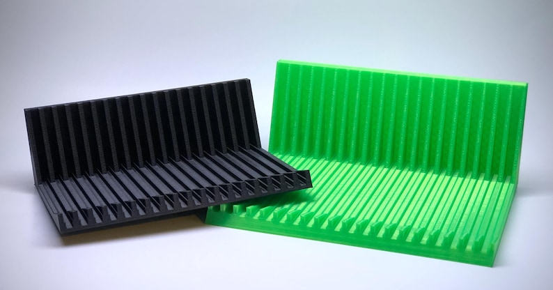 May include: Two 3D printed corner shelves, one black and one green. The shelves have a ridged design and are angled to create a corner display.