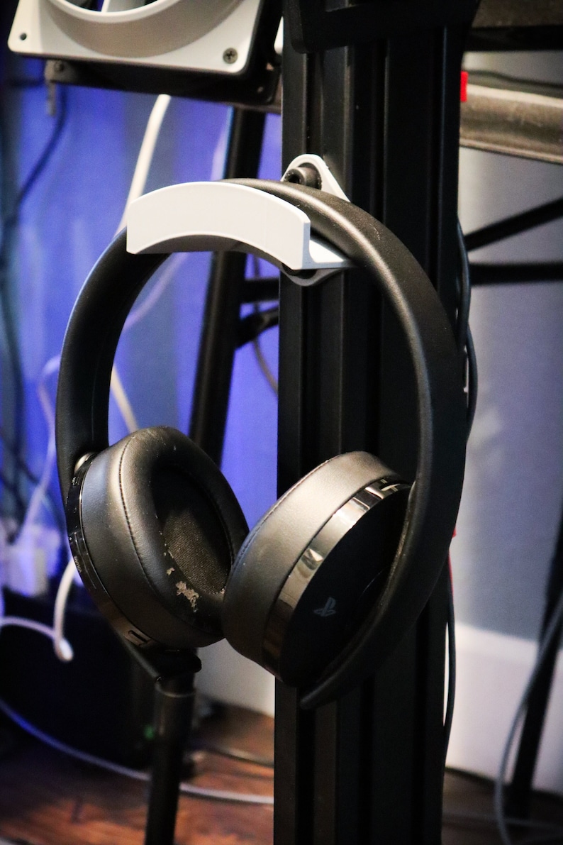 May include: A black headset with a PlayStation logo hangs on a black metal stand with a white plastic hook. The headset is hanging by the headband.