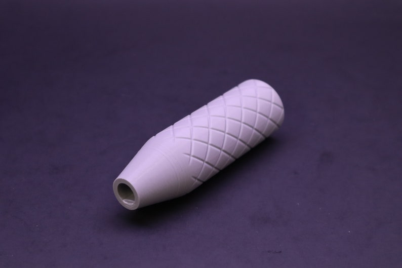 May include: A light grey, cylindrical object featuring a diamond pattern carved into its surface. The object narrows to a smaller opening at one end. It appears to be a handle or grip for a tool or device.