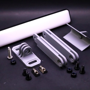 Może przedstawiać: Zestaw jasnoszarych elementów montażowych do kamery, w tym wsporniki, śruby i długie, prostokątne światło. Komponenty są ułożone na ciemnej powierzchni, sugerując motyw techniczny lub fotograficzny.