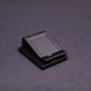 May include: A black metal clip, likely for holding papers or other items. The clip has a rectangular shape with a hinged top and a small hole. The object is set against a dark gray background.