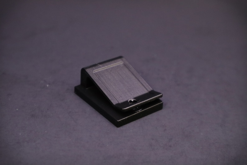 May include: A black metal clip, likely for holding papers or other items. The clip has a rectangular shape with a hinged top and a small hole. The object is set against a dark gray background.