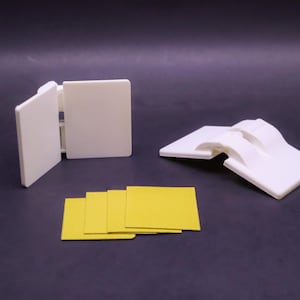 May include: White plastic hinges and yellow square pads are displayed against a dark background. One hinge is open, another is closed, and a third is partially open. The yellow pads are stacked in a neat pile.