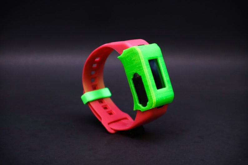 Pode incluir: Uma pulseira vermelha e verde com um visor digital. A pulseira &eacute; feita de silicone e tem uma capa de pl&aacute;stico verde sobre o visor.