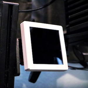 May include: A square digital display screen featuring a solid white frame and a dark, reflective glass panel. The device is mounted on a black vertical support structure, positioned at an angle to show the depth of the white plastic casing.