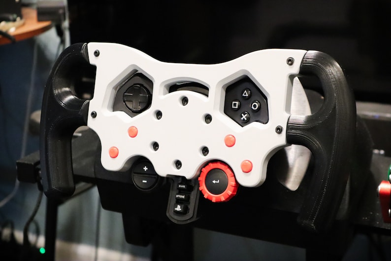 May include: A white and black racing steering wheel with red buttons and a black and red knob. The steering wheel has a variety of buttons and controls, including a D-pad, buttons for the PlayStation console, and a knob for adjusting settings.