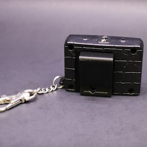 May include: Black rectangular keychain accessory with a silver-colored chain and clasp. The accessory has a textured surface and a black clip on the back. The keychain is designed to attach to keys or other items.