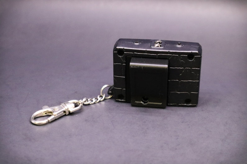 May include: Black rectangular keychain accessory with a silver-colored chain and clasp. The accessory has a textured surface and a black clip on the back. The keychain is designed to attach to keys or other items.