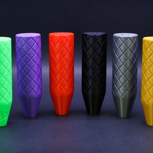 May include: Six shift knobs in a row, each with a diamond pattern. The colours are green, purple, red, black, grey, and gold. The knobs have a cylindrical top and a tapered base, designed for a car's gear stick.