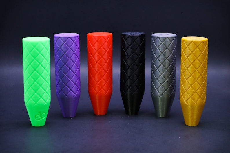 May include: Six shift knobs in a row, each with a diamond pattern. The colours are green, purple, red, black, grey, and gold. The knobs have a cylindrical top and a tapered base, designed for a car's gear stick.