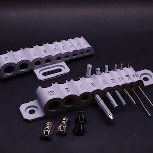 May include: A white plastic tool holder with slots for holding various sizes of screwdrivers. The holder has numbers 10, 8, 6, 5, 4, 3, 2, 1, and 1/2 printed on the slots. The holder is sitting on a black surface with a set of screwdrivers and two silver metal fasteners.