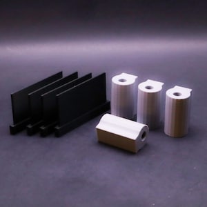 May include: An assortment of metal components, including four black rectangular pieces and four cylindrical pieces with a hole at the top. One of the cylindrical pieces is rectangular. The items are arranged on a dark surface.