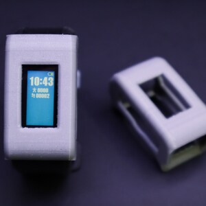 May include: A white and gray fitness tracker with a blue screen displaying the time 10:43. The tracker has a black band and is sitting on a dark gray surface. There is a second white and gray object in the background.