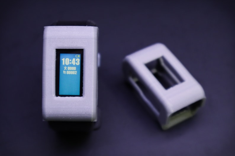 May include: A white and gray fitness tracker with a blue screen displaying the time 10:43. The tracker has a black band and is sitting on a dark gray surface. There is a second white and gray object in the background.