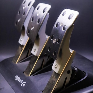 May include: A set of silver and black racing pedals with the Logitech G logo. The pedals have a metal finish with multiple holes and are mounted on a black base. The pedals are designed for use with racing simulation games.