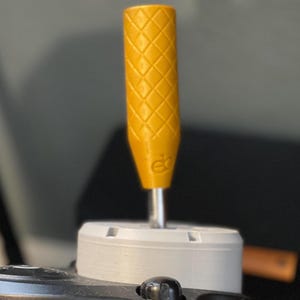 May include: A yellow screwdriver handle with a diamond pattern grip. The handle is attached to a silver metal shaft and a white base. The handle has a small logo at the base. The background is blurred.