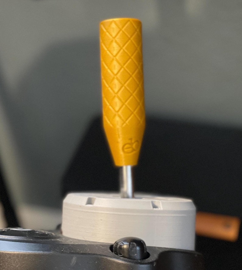 May include: A yellow screwdriver handle with a diamond pattern grip. The handle is attached to a silver metal shaft and a white base. The handle has a small logo at the base. The background is blurred.