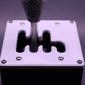 May include: A white plastic gear shifter with a black base and a silver gear shift knob. The gear shifter has a cutout for the gear pattern, which is visible in the image.