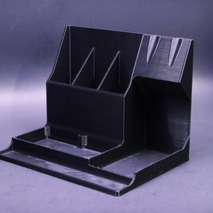 May include: A black plastic desk organizer with multiple compartments. The organizer has a tall section with three vertical slots, a wide shallow tray, and a small open compartment.
