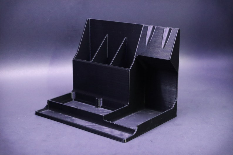 May include: A black plastic desk organizer with multiple compartments. The organizer has a tall section with three vertical slots, a wide shallow tray, and a small open compartment.