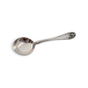 May include: A silver-toned metal spoon featuring a round bowl and a handle decorated with an ornate, raised floral scroll pattern. The polished surface of the bowl reflects light, showing a smooth, curved interior and a detailed decorative stem.