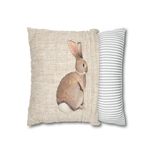 May include: A decorative throw pillow with a brown rabbit illustration on a beige linen fabric. The back of the pillow is a gray and white striped fabric.