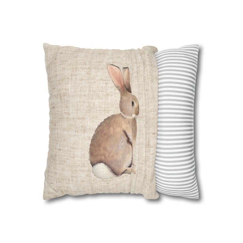 May include: A decorative throw pillow with a brown rabbit illustration on a beige linen fabric. The back of the pillow is a gray and white striped fabric.
