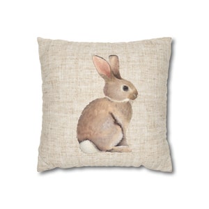May include: A beige throw pillow with a watercolor illustration of a brown rabbit sitting on a white background.