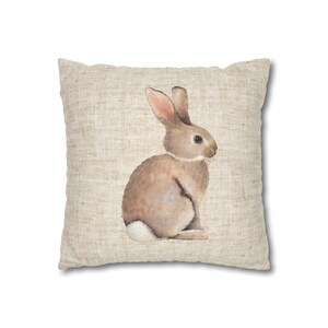 May include: A brown rabbit with pink ears sits on a beige linen pillow. The rabbit is facing to the right and has a white tail.