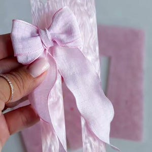 May include: A light pink fabric bow attached to a rectangular, marbled light pink acrylic bookmark. The bookmark features the text LILACPEACHES handmade with a small floral and bow graphic at the top and a star-shaped cutout near the center.